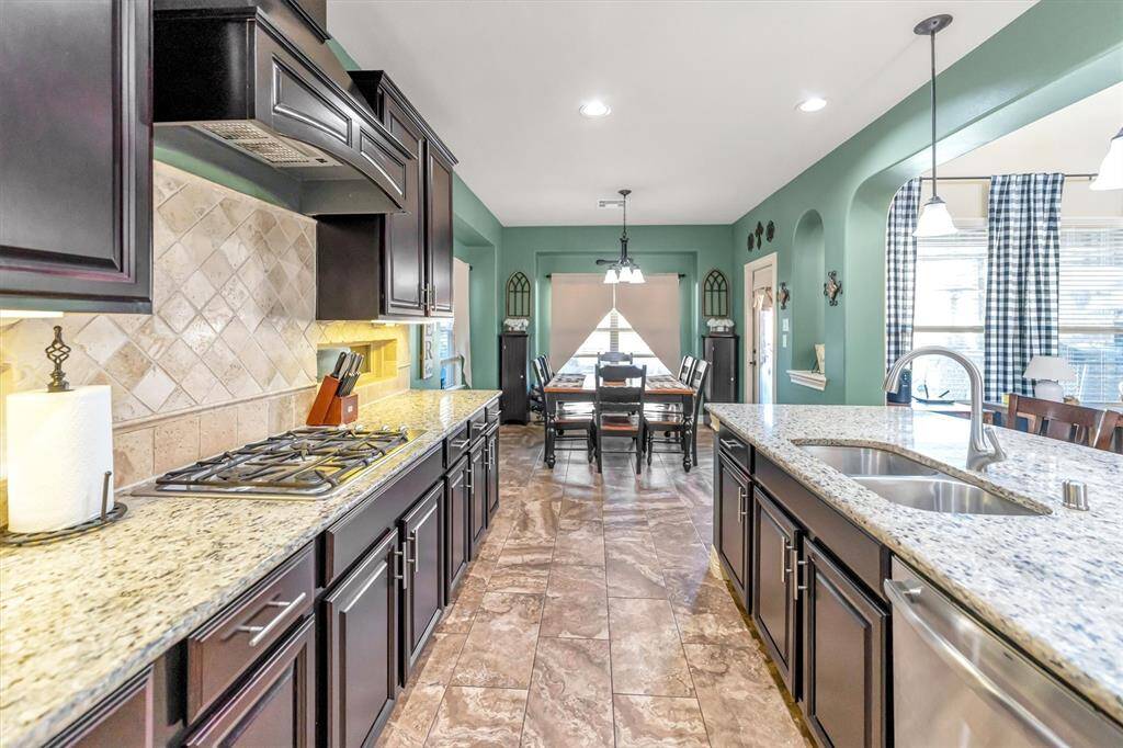Explore this stunning modern kitchen featuring dark wood cabinets, granite countertops, and a gas stove. The spacious island with a sink and pendant lighting seamlessly connects to a sunlit dining area with elegant tile flooring.