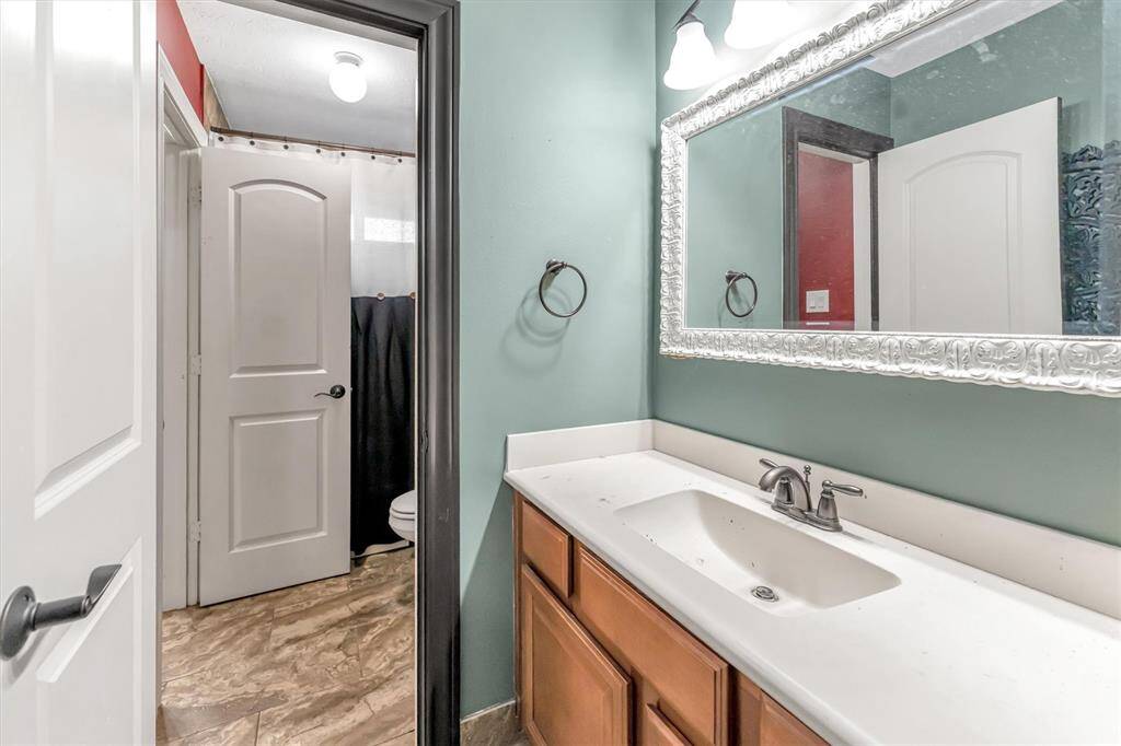 Discover a stylish, well-lit modern bathroom featuring a large framed mirror, sleek single-sink vanity with wooden cabinetry, and soft, neutral walls. A doorway leads to a private shower area with a curtain.