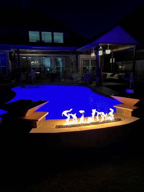 Evening view of the pool area highlighting the poolside fireplace. Notice the pendant lighting above the outdoor seating area.
