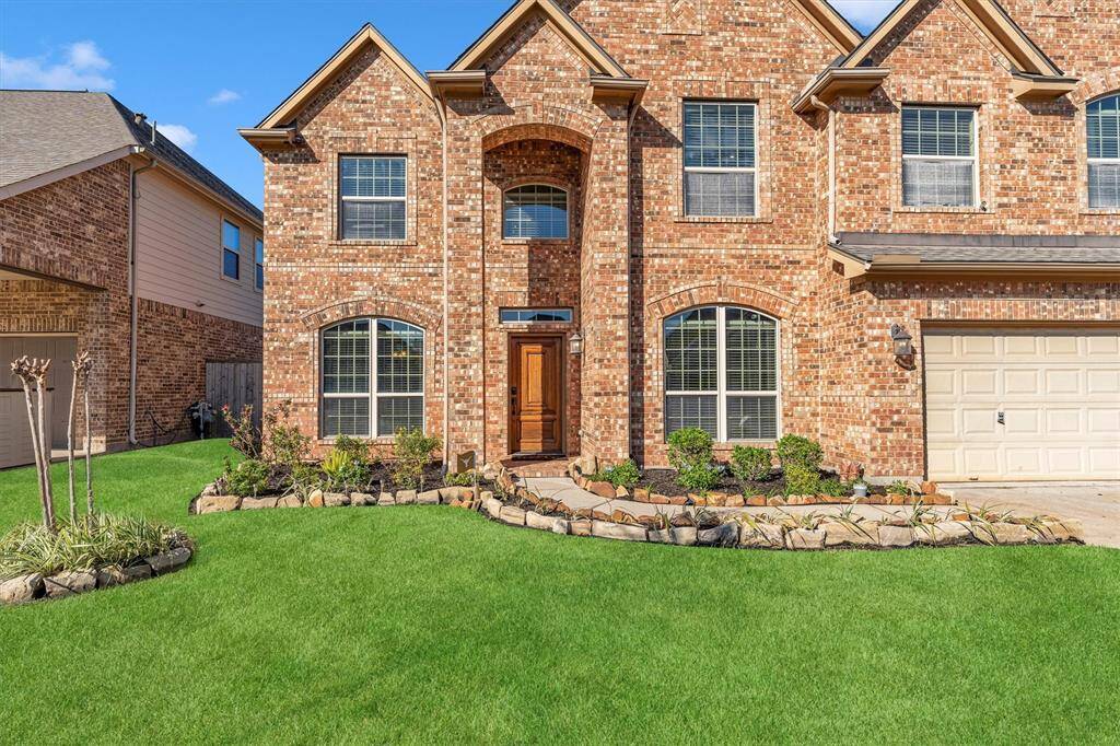 Charming two-story brick home with a lush lawn, elegant landscaping, large windows for natural light, and a spacious attached garage.