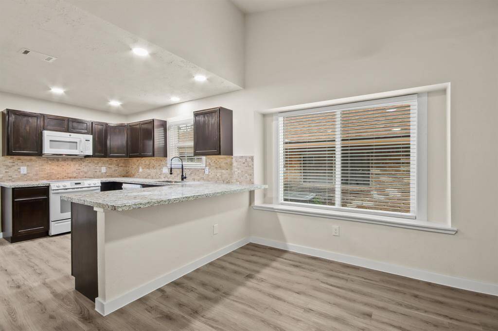 This modern kitchen features dark cabinetry, granite countertops, stainless steel appliances, and a stylish backsplash. The open layout includes a large window for natural light and a spacious dining area with wood flooring.