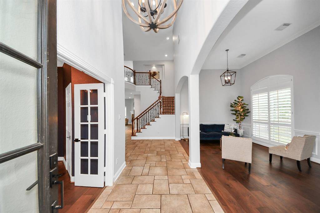 Spacious entryway with high ceilings, featuring a mix of tile and wood flooring. The open layout includes a staircase with wrought iron railing, a cozy seating area with large windows, and elegant lighting fixtures.