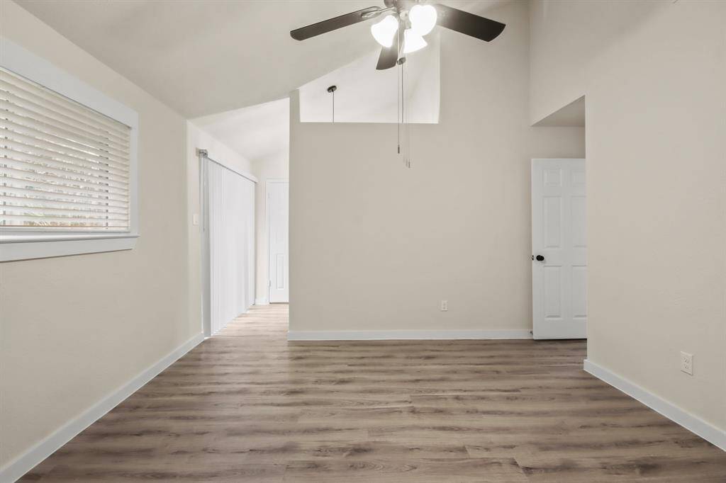 This room features high ceilings with a ceiling fan, neutral walls, and wood-look flooring. It has ample natural light from a window with blinds and offers a clean, open space with a white door leading to another room.