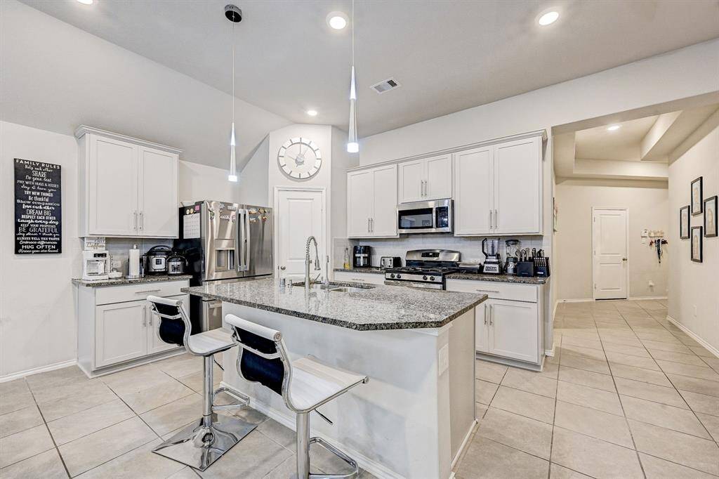 This modern kitchen features sleek white cabinetry, stainless steel appliances, and a spacious granite island with seating. Pendant lighting and ample counter space make it ideal for cooking and entertaining. The open layout connects seamlessly to the adjacent areas.