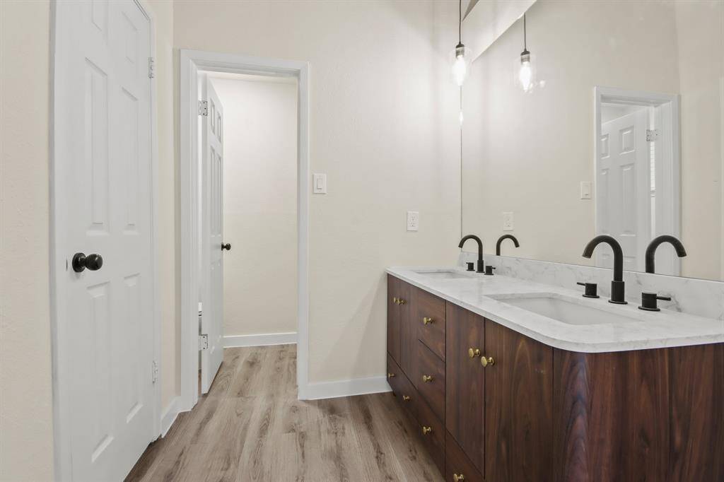 This bathroom features a modern double vanity with sleek black fixtures, marble countertops, elegant pendant lighting, and wood-grain cabinetry, complemented by light-colored walls and flooring.
