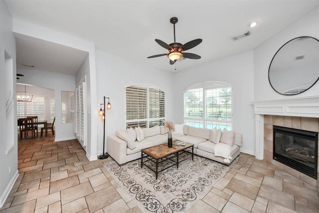 This inviting living room features a cozy fireplace, large windows with plantation shutters, and a stylish ceiling fan. The open layout flows into the dining area, and the neutral tones complement the elegant tile flooring, creating a warm and welcoming space.