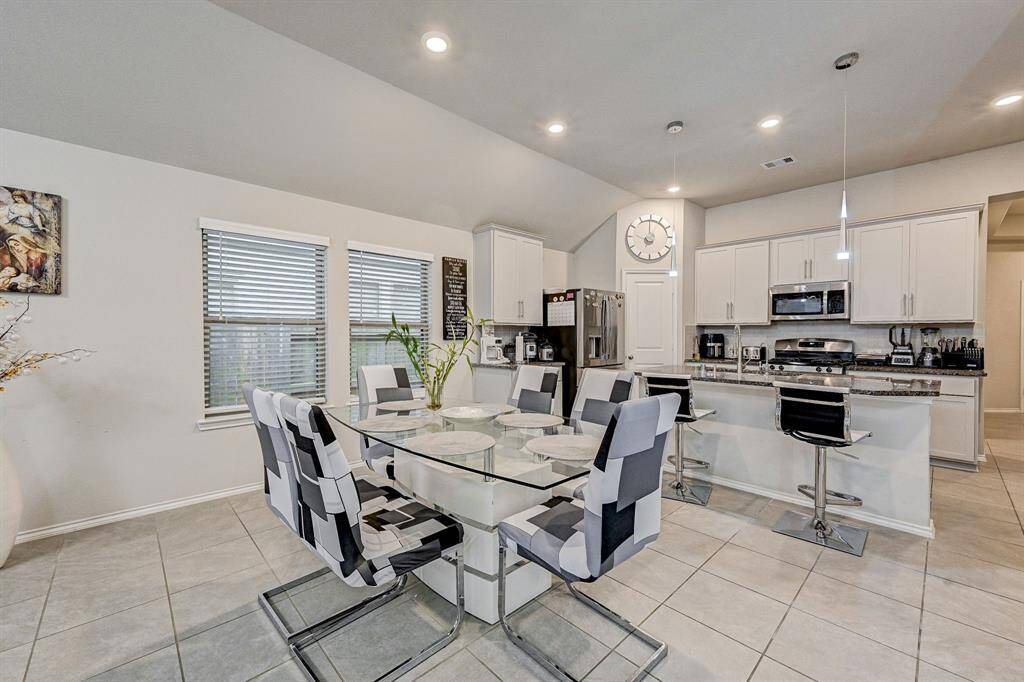 This photo showcases a modern kitchen and dining area with sleek tile flooring, a glass-top dining table, and stylish chairs. The kitchen features white cabinetry, stainless steel appliances, and a breakfast bar with pendant lighting, creating a bright and inviting space.