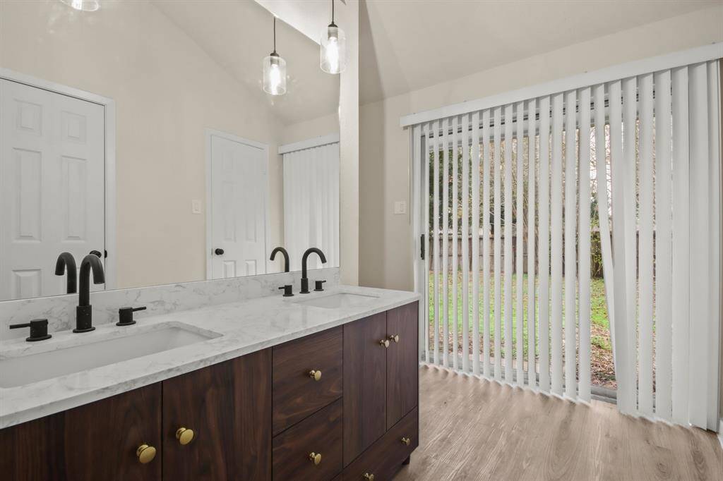 This bathroom features a double sink vanity with marble countertops, modern black fixtures, and elegant pendant lighting. A sliding glass door with vertical blinds opens to a view of the backyard, enhancing natural light.