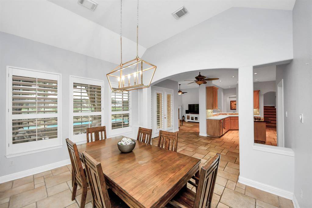 Bright dining area featuring a modern pendant light. It opens to a spacious kitchen and living room with tile flooring, large windows with plantation shutters, and a view of the sun room and backyard pool area.