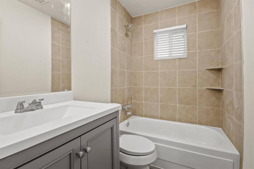 This bathroom features a modern design with beige tiled walls, a white bathtub, a sleek gray vanity with a white countertop, and a window with blinds for natural light.