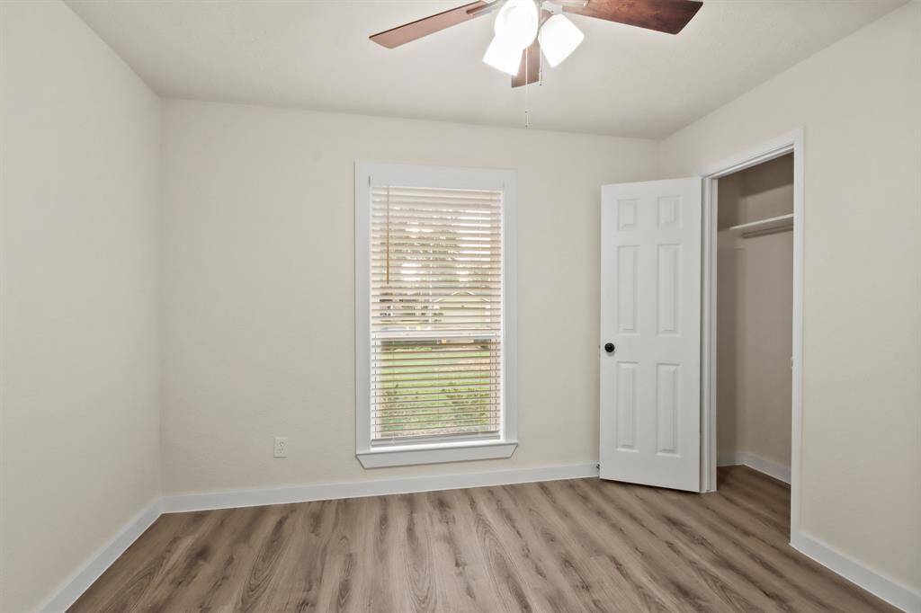 This is a bright, small room with neutral walls and wood-style flooring. It features a window with blinds, a ceiling fan with lights, and an open closet with a white door.
