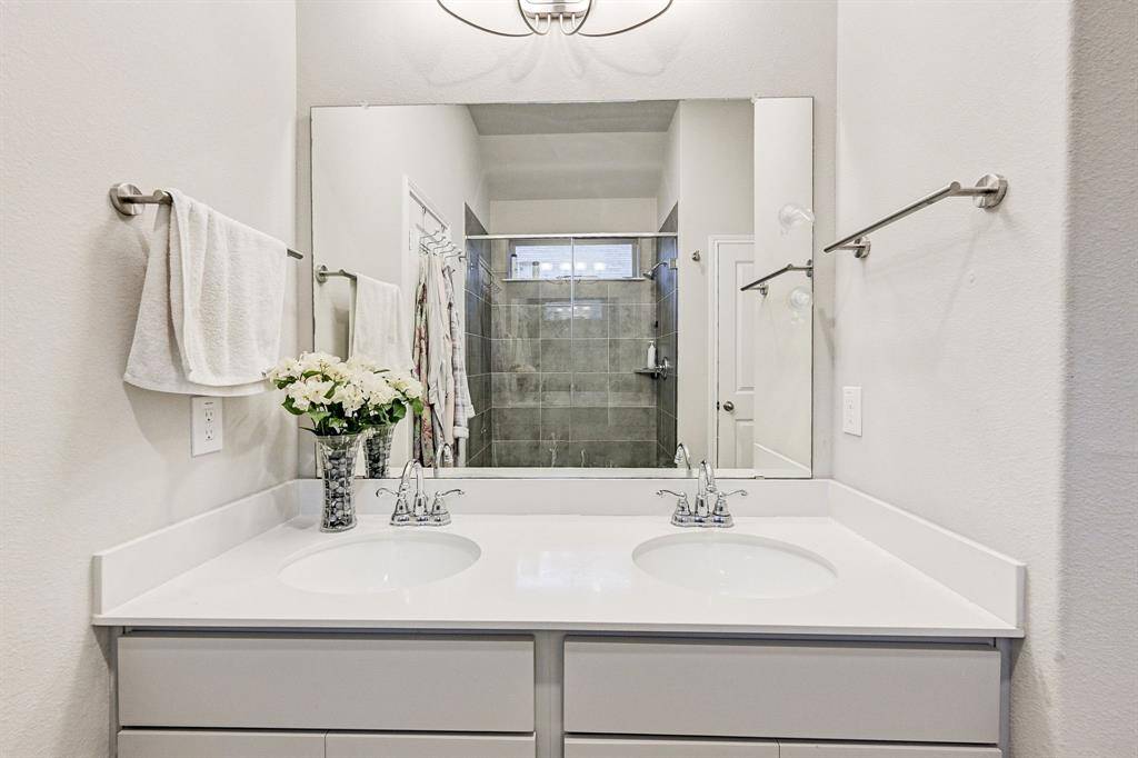 This photo showcases a modern bathroom with a double vanity featuring sleek white countertops and chrome fixtures. A large mirror expands the space, and there's a spacious shower with gray tiles. The light walls enhance the overall brightness of the room.