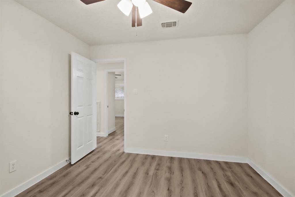 This room features neutral walls, wood-like flooring, a ceiling fan with lights, and an open door leading to another space, creating a bright and inviting atmosphere.