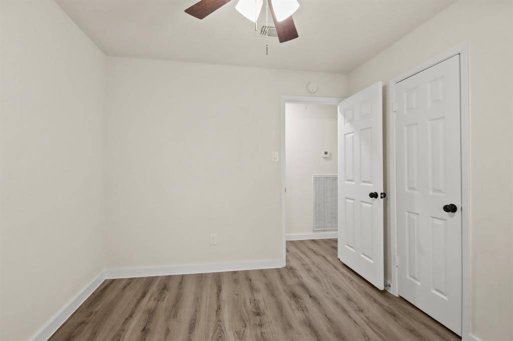 This photo shows a small, bright room with light walls and wood-style flooring. It features a ceiling fan and two white doors, one leading to an adjacent area.