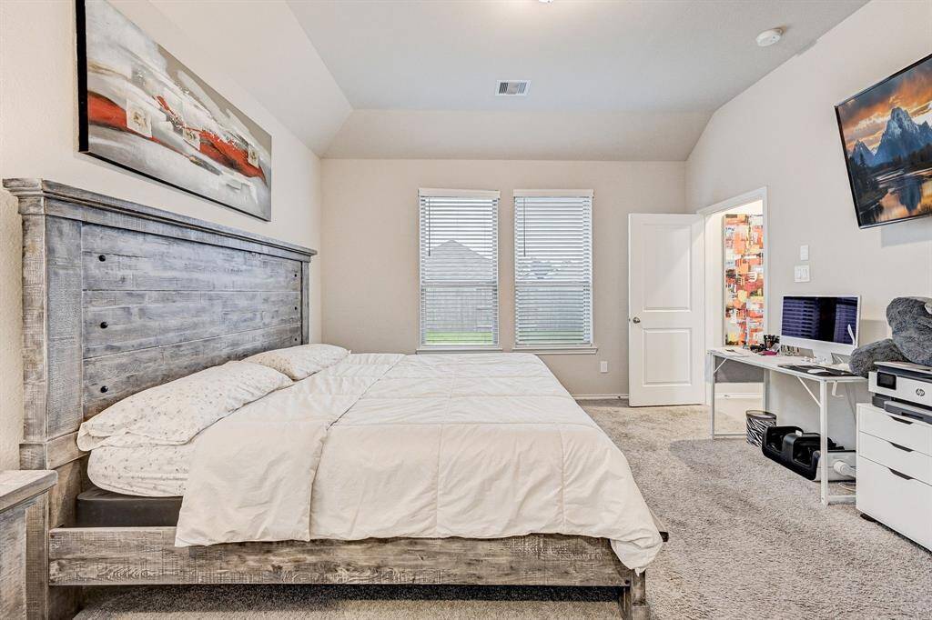 This photo showcases a spacious bedroom with a rustic wooden bed, neutral walls, and carpeted flooring. Natural light streams through two large windows. There's a wall-mounted TV and a desk with a computer setup, providing a comfortable work-from-home space. An open door leads to an attached bathroom, adding convenience.