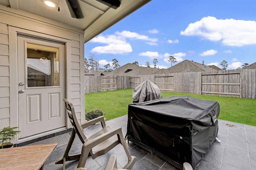 This photo showcases a cozy, covered patio area with seating and a grill, overlooking a private, fenced backyard. The space is ideal for outdoor relaxation and entertaining, with a view of neighboring rooftops under a clear blue sky.