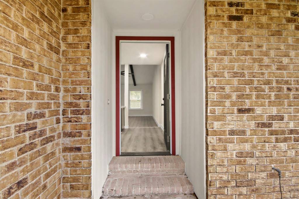 This photo shows the entrance of a home with a brick exterior and a welcoming front door. Inside, there is a glimpse of an open, bright interior with light flooring and a visible window, suggesting a spacious living area.