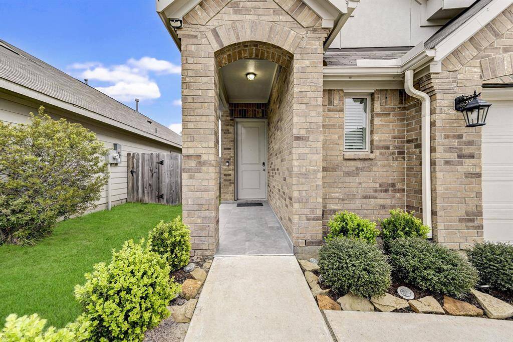 This photo showcases a welcoming entrance to a brick home, featuring a neatly landscaped front yard with shrubs and a small lawn. The arched entryway leads to a covered porch and front door, offering a charming and tidy curb appeal.