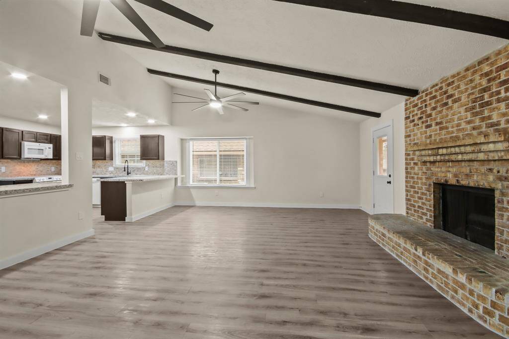 Spacious living area with high ceilings and exposed beams, featuring a cozy brick fireplace. The room flows into an open kitchen with modern cabinetry and a large window. Fresh flooring and neutral tones create a welcoming atmosphere.