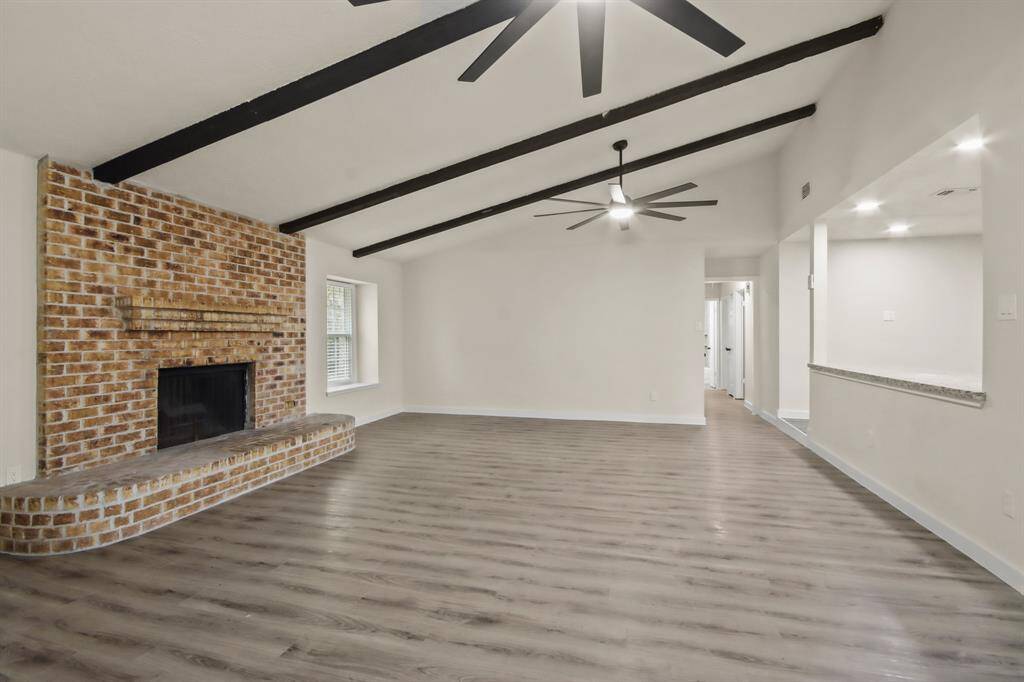 This spacious living room features a cozy brick fireplace, vaulted ceilings with exposed beams, modern ceiling fans, and sleek wood flooring, offering a warm and inviting atmosphere.