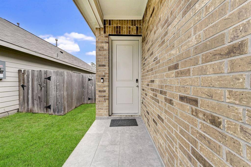 This photo shows a welcoming entryway with a brick exterior wall, a neatly paved walkway, and a wooden fence leading to a side yard with green grass.