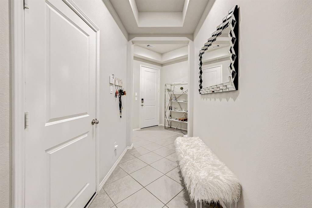 This photo shows a modern, well-lit entryway with neutral tones. It features a tiled floor, a stylish mirror, and a cozy bench. A door leads to the interior, and there's a shelving unit for storage or decor.