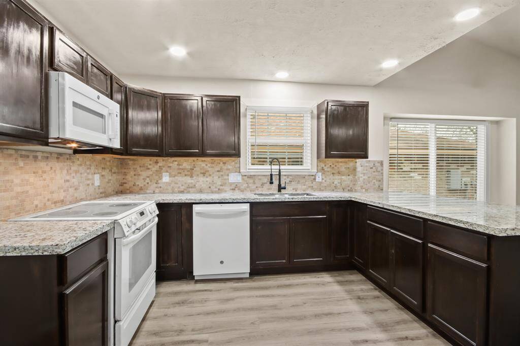 This kitchen features dark wood cabinets, granite countertops, a tile backsplash, and modern appliances. It has ample natural light from a window above the sink, offering a spacious and inviting atmosphere.