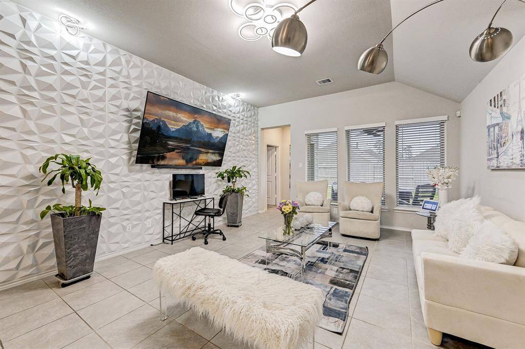 This modern living room features a stylish 3D accent wall, large TV, and contemporary lighting. It has a cozy seating area with plush cushions, a glass coffee table, and elegant decor. Large windows provide natural light, enhancing the spacious feel.