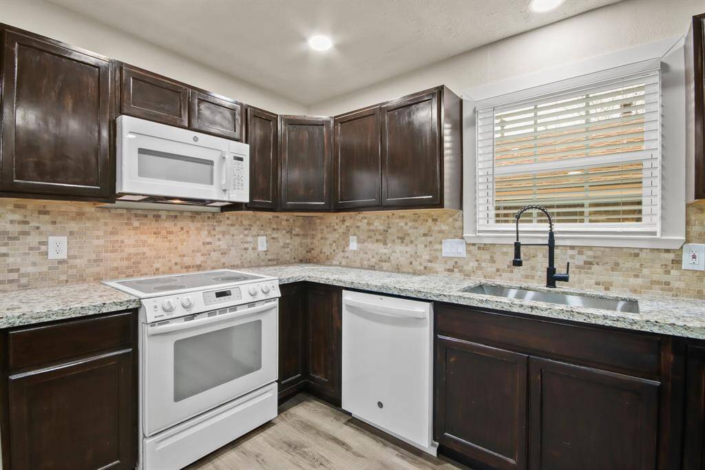 This kitchen features dark wood cabinets, granite countertops, a tile backsplash, and modern white appliances, including a built-in microwave and dishwasher. A large window above the sink provides natural light.