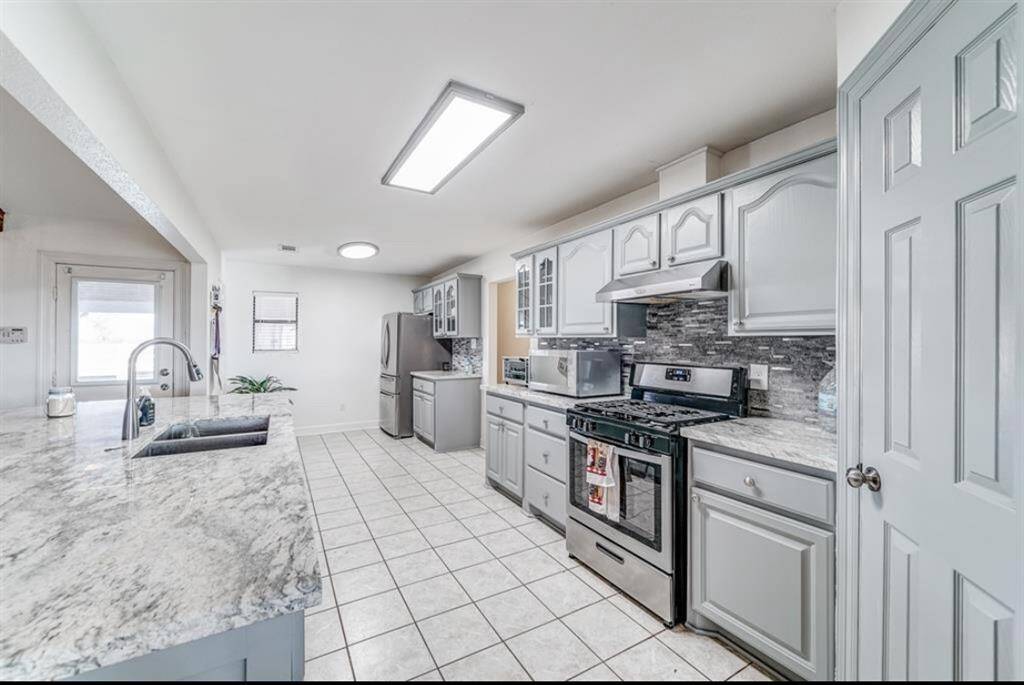 This kitchen features sleek gray cabinetry, modern stainless steel appliances, and a spacious island with a marble countertop. The tiled floor and ample natural light create a bright and inviting space.