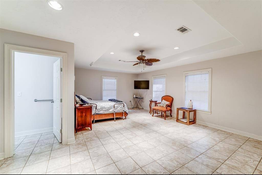 Spacious bedroom with tiled flooring and neutral walls. Features a ceiling fan, ample natural light from two windows, and a cozy seating area. Adjacent bathroom for convenience.