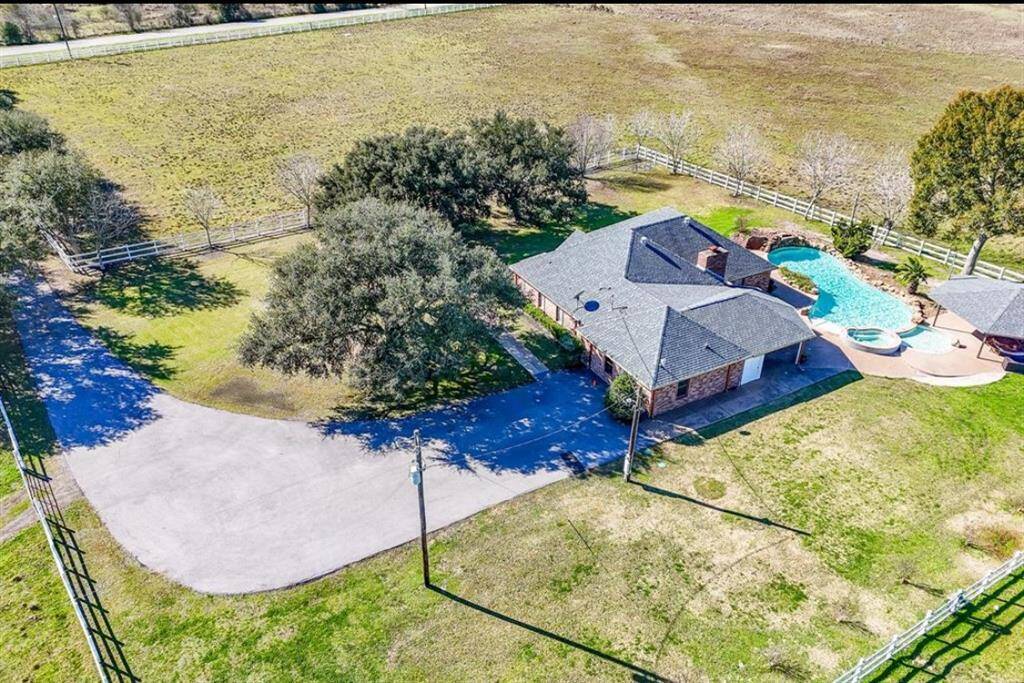 This aerial view showcases a spacious, single-story brick home with a large yard, featuring a swimming pool and patio area. The property is surrounded by a white fence and mature trees, offering privacy and a serene rural setting.