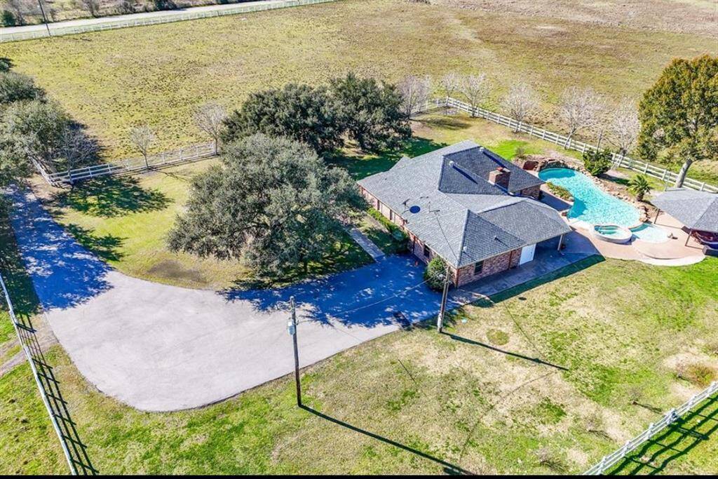 This aerial view showcases a charming brick home on a spacious, fenced property with a large driveway, mature trees, and a backyard featuring a pool and hot tub. Ideal for privacy and outdoor enjoyment.