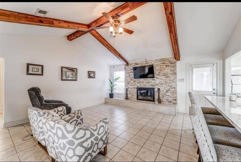 This cozy living room features a vaulted ceiling with exposed wooden beams, a stone fireplace, and tiled floors. Ample natural light fills the space, complemented by a ceiling fan and modern furnishings. Ideal for relaxation and entertaining.