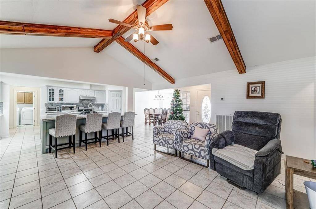 This spacious living area features a high ceiling with exposed wooden beams and a ceiling fan. The open layout connects to a modern kitchen with a large island and bar seating. Adjacent is a dining area and a cozy seating space, all set on tiled flooring. The room is bright and inviting, perfect for family gatherings.