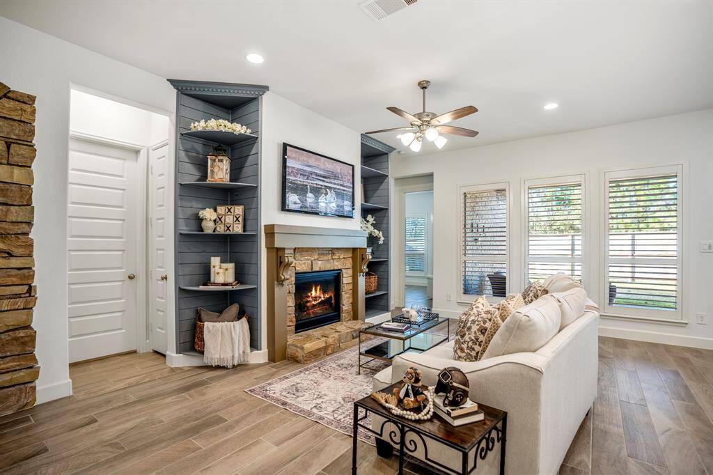 This living room is a warm and inviting space, centered around a stunning fireplace. Symmetrical built-in shelves flank the fireplace, painted in a contrasting muted blue-gray tone. The shelves offer ample space for decorative accents, books, and personal touches, enhancing the room's cozy feel. Large windows with plantation shutters let in natural light and offer views of the outdoor space, maintaining a bright and airy atmosphere.