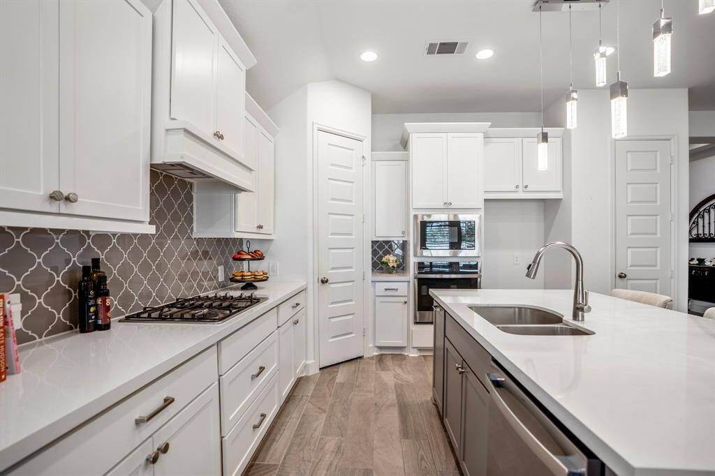 This kitchen design features white cabinetry and clean lines. White cabinets for  sleek and bright look with simple hardware. Quartz countertops along with the geometric-patterned backsplash in neutral tones adds a subtle yet stylish element provide an elegant touch. A stainless steel cooktop embedded seamlessly into the counter and the built-In oven and microwave  appliances integrated into the cabinetry give a streamlined appearance.