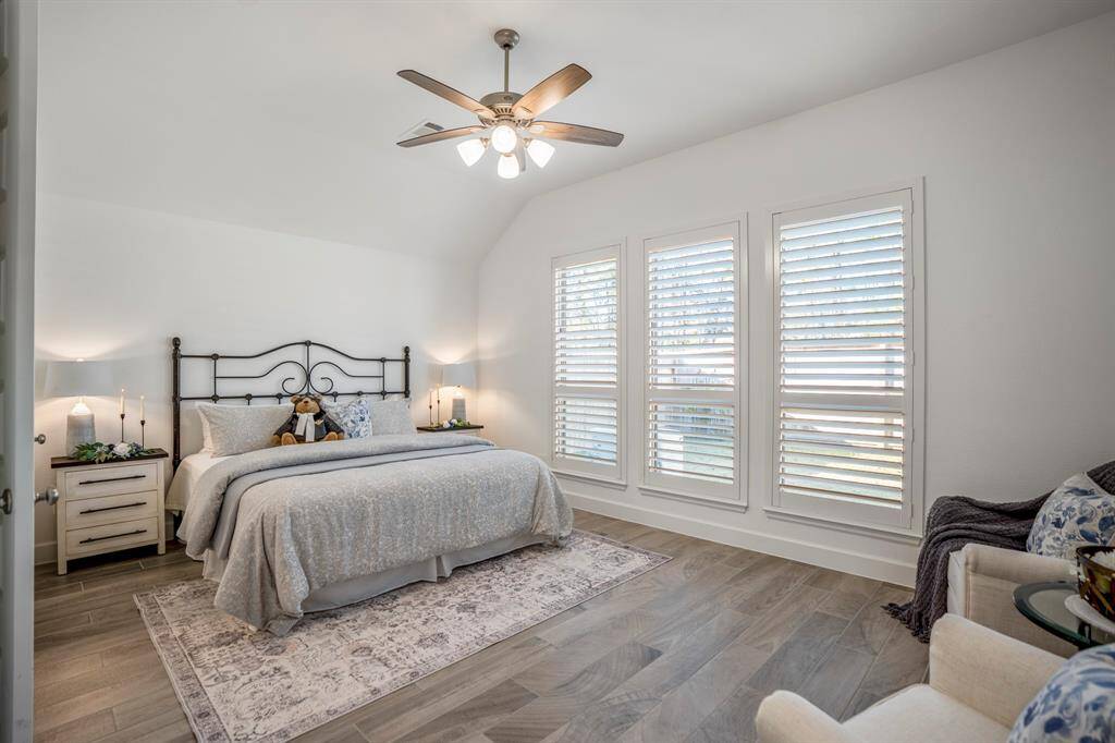 A plush bed with soft gray bedding and decorative pillows creates a cozy and relaxing retreat. A row of large windows with plantation shutters allows natural light to fill the room while offering privacy when needed. This primary bedroom exudes comfort and charm, making it a perfect retreat for rest and relaxation.