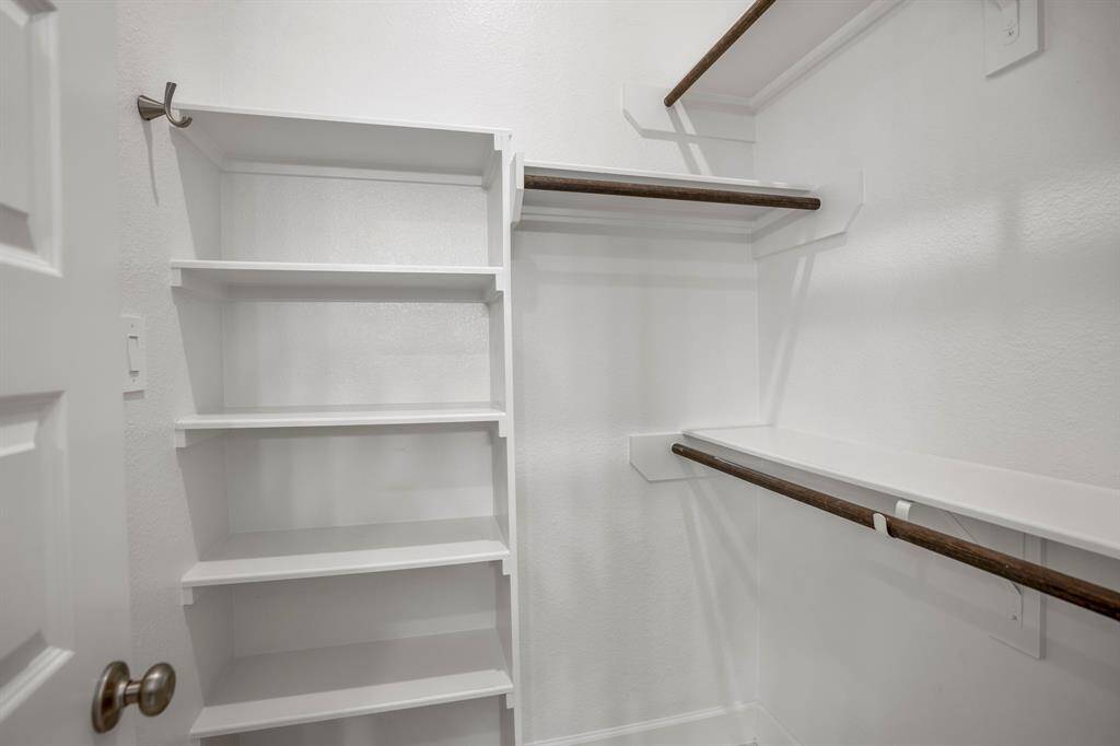 This is one of the two walk-in closets in the primary suite, designed to maximize storage and organization. Multiple built-in shelves offer space for folded clothes, shoes, or accessories. Crisp white walls and shelves create a cohesive and clean aesthetic, making the space feel larger and brighter. With two walk-in closets in the primary suite, this design offers ample storage to accommodate any wardrobe while maintaining an organized and functional set up.