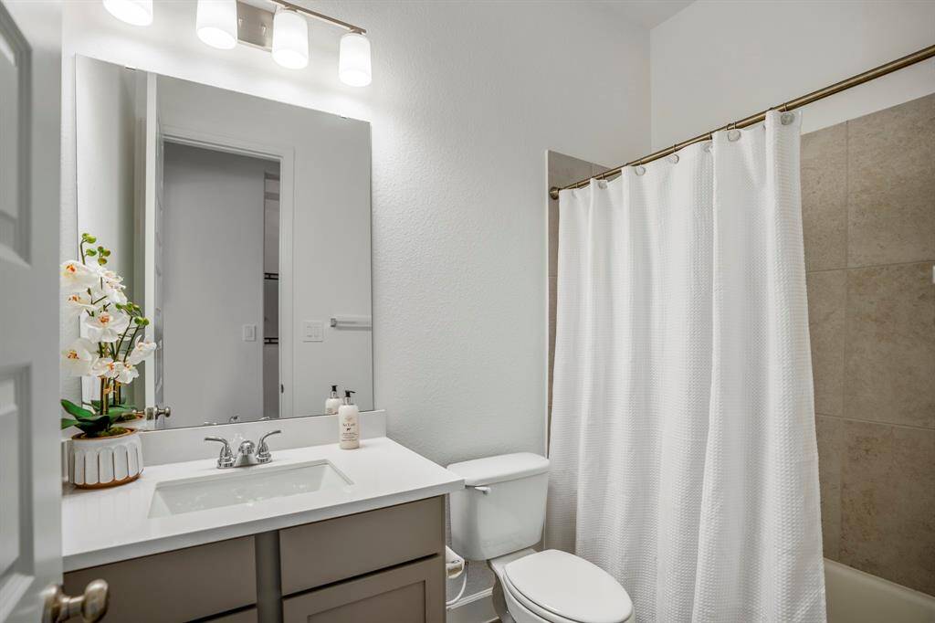 This secondary bathroom is both functional and visually appealing, offering a versatile space for family members or guests. A single vanity with a sleek white countertop and undermount sink .Gray cabinetry provides storage while complementing the neutral palette. A large mirror above the vanity enhances the sense of space, illuminated by a modern three-light fixture with frosted glass shades. A standard bathtub with a tiled surround in neutral tones for a clean and timeless look.