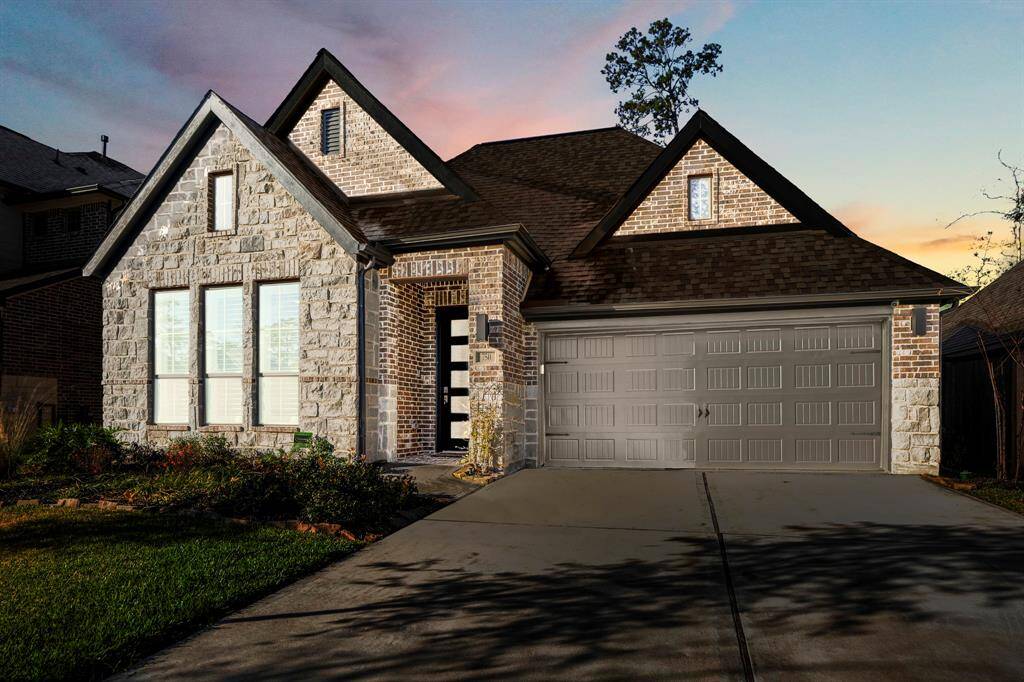 This beautiful single-story house with a mix of stone and brick exterior. It features a gable roof with dark shingles and a large two-car garage with a paneled design in a muted tone.