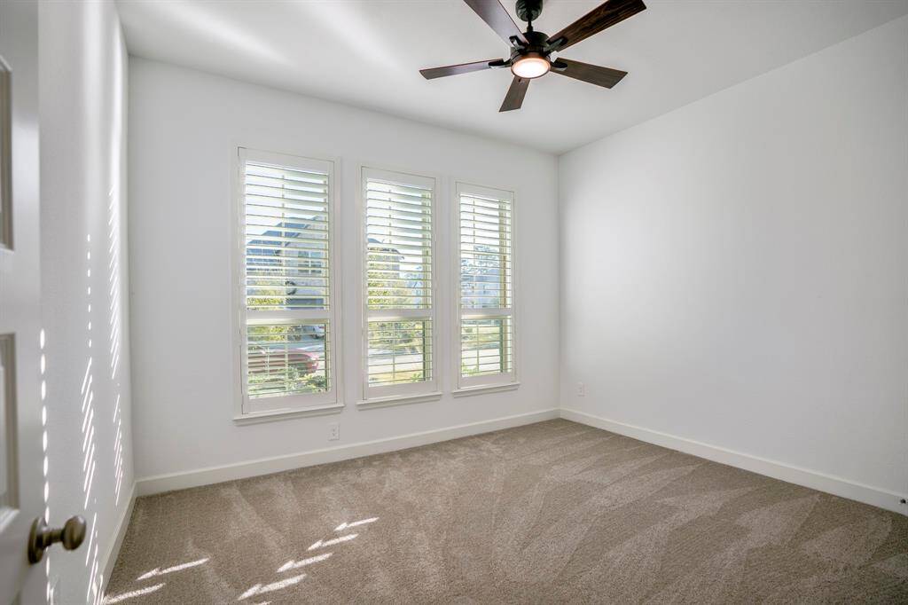 This secondary bedroom provides a comfortable and adaptable area, ideal for family members or guests. A row of three large windows with plantation shutters allows abundant natural light while maintaining privacy. The windows provide a view of the outdoor surroundings, enhancing the room's airy feel. Soft, neutral-toned carpeting covers the floor, adding warmth and comfort to the space. The room offers a balanced layout with enough space to accommodate a bed, furniture, or other desired elements.