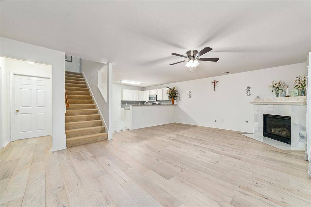 This is a spacious living area with light engineer hardwood flooring, a modern ceiling fan, a fireplace, and an open-concept layout leading into the kitchen. The staircase provides access to an upper level, and there's a white doorway that likely leads to Primary bedroom and half bath