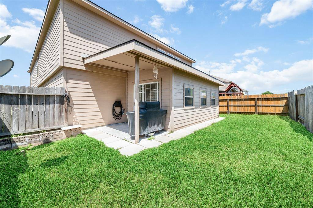 This is a well-maintained backyard with a lush green lawn, featuring a covered patio area and a high privacy fence. The exterior of the house shows a clean, modern design with neutral colors.