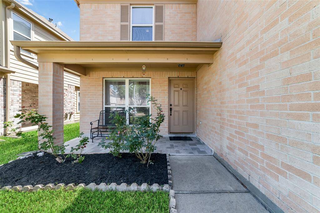 This is a cozy, covered front porch area of a brick home, featuring a neutral-toned front door, tiled walkway, and landscaping with fresh mulch.
