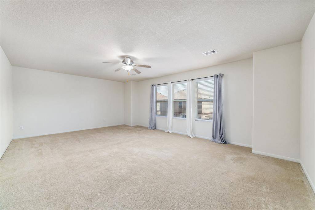 You're looking at a spacious, well-lit room with neutral carpeting, fresh white walls, and a ceiling fan. It features ample natural light from three large windows dressed with elegant gray curtains.