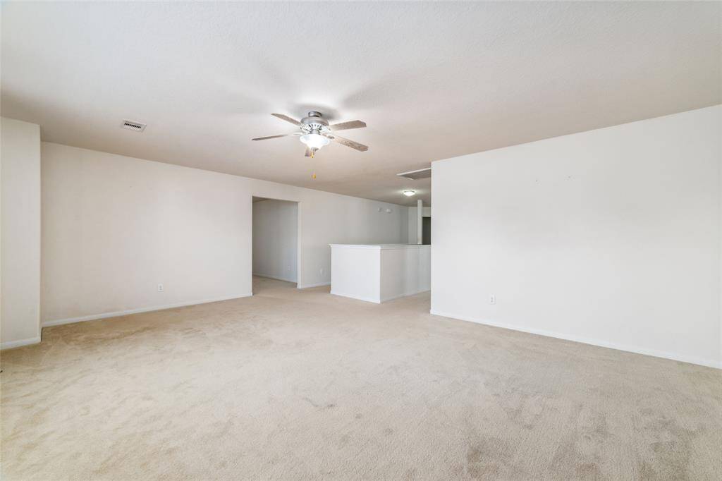This is a spacious, well-lit room with neutral walls and carpeting, featuring a ceiling fan and an open layout leading into another room, offering a blank canvas for personalization.