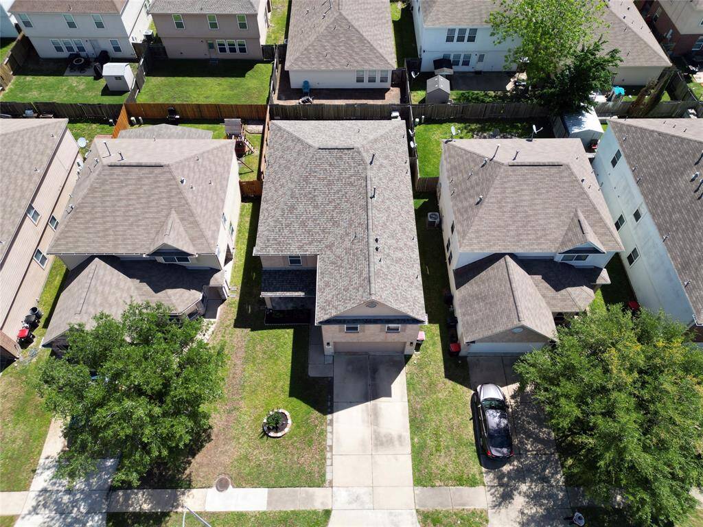 This aerial photo shows a residential neighborhood with closely spaced, two-story homes featuring gray shingle roofs, small backyards with privacy fences, and an unobstructed front driveway. A mature tree is visible in the front yard, adding a touch of greenery to the property.