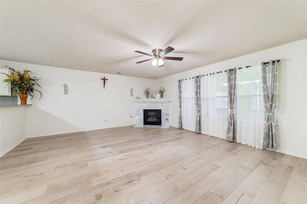 This is a spacious and well-lit living room featuring contemporary engineered hardwood flooring, a cozy fireplace, large windows with stylish curtains, and a high ceiling with a modern fan. The neutral color palette offers a blank canvas for personal decor preferences.