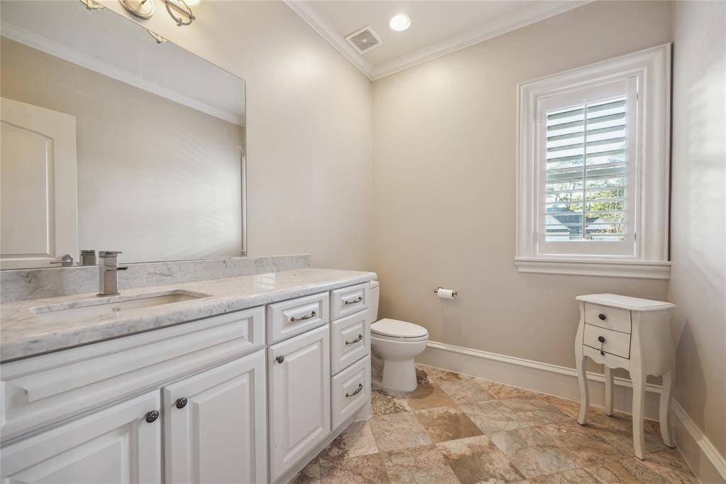 This elegant bathroom features a spacious stone countertop, pristine white cabinetry, and a large mirror. Natural light flows through the window with plantation shutters, highlighting the stylish tile flooring.
